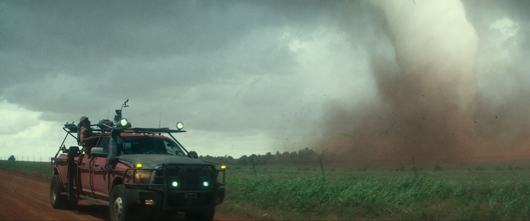 A vertical still from the movie 'Twisters' shows a red truck on the right equipped with tornado chasing hear and extra lights. A man leans halfway out the window to look towards a nearby tornado on the right side of the image.