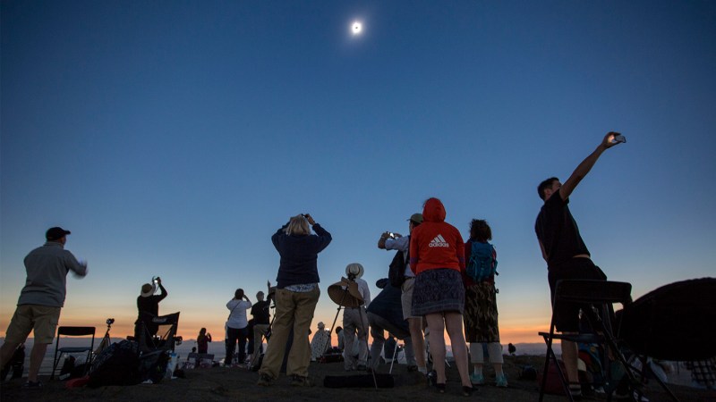 About a dozen people with their backs turned to the camera watch the August 21, 2017 eclipse. The people are looking up into the sky at a white sun with a black dot in the center. The sun is at the top center of the image. The sky is dark blue gradually darkening to midnight at the upper corners of the picture. The horizon is yellow and orange as if at sunset. Several people are taking pictures with cell phones held aloft. One person at the center right in the back of the group wears a red hooded sweatshirt with an Adidas logo on the back and a skirt.